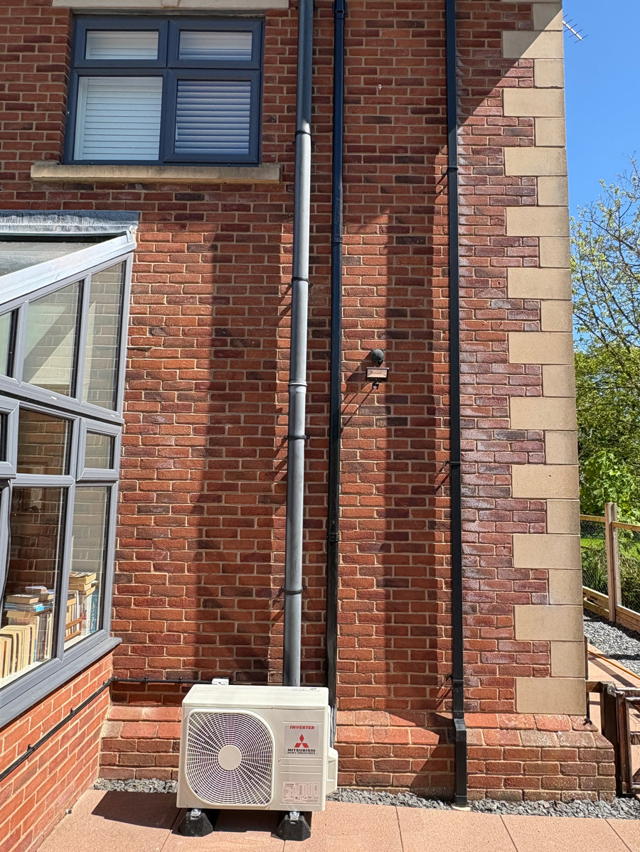 Outdoor Mitsubishi Heavy Industries air conditioning unit on feet beside a brick wall, with vertical pipework